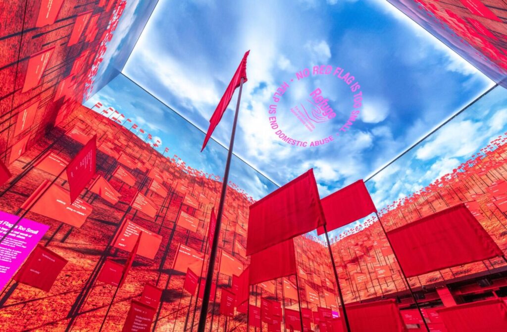 Looking up to the ceiling of the Outernet, London. The screen walls are filled with red flags up to a screen of a blue sky with clouds and the Refuge logo on the ceiling. In the foreground are physical red flags on poles