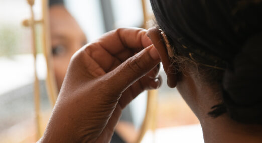 Close-up of a Black woman putting a hearing aid into her left ear. She is dressed in all black, with her pulled back by a scarf, and looking into a tabletop mirror.