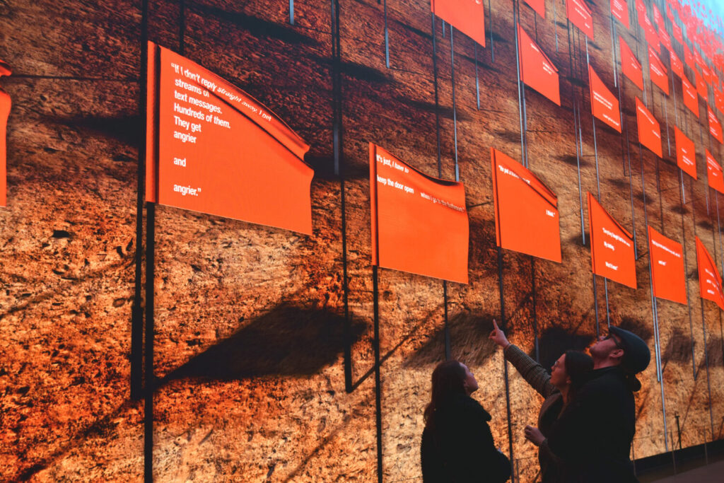 Three people standing in front of a large screen filled with red flags , looking and pointing