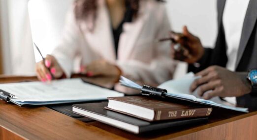 In the foreground is a book titled 'The Law'. Out of focus behind it, a man and a woman's hands can be seen with legal documents.