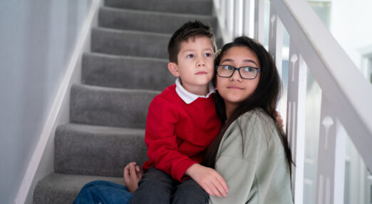 Photo of two children sat together on carpeted stairs. The girl is wearing glasses and a grey jumper, the boy is sat on her lap in red school uniform. They are both unsmiling, and looking off to the right of the camera.
